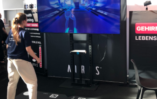 Frau steht vor großem Bildschirm und spielt ein Bewegungsspiel in einem Messestand mit schwarzem Hintergrund und weißen Markierungen auf dem Boden