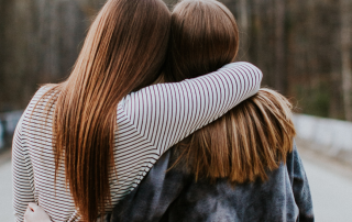 Zwei Frauen mit langen Haaren umarmen sich von hinten auf einem Waldweg.
