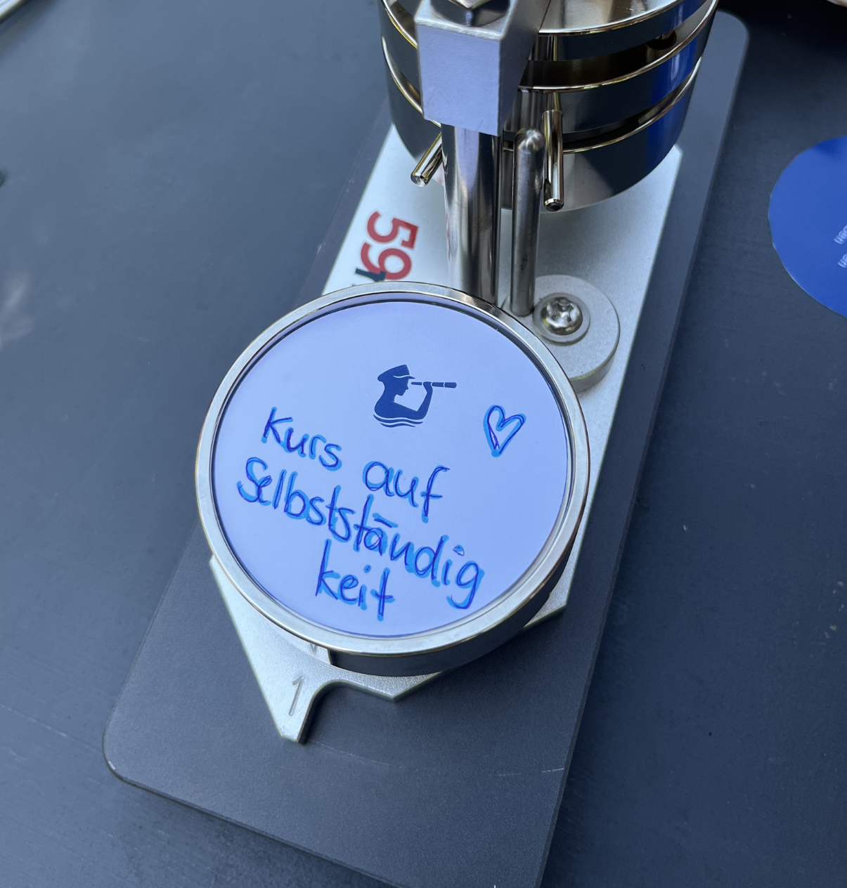 Nahaufnahme eines runden weißen Aufklebers mit blauer Handschrift 'kurs auf selbstständigkeit' und einem kleinen Herzsymbol in einer Buttonmaschine.