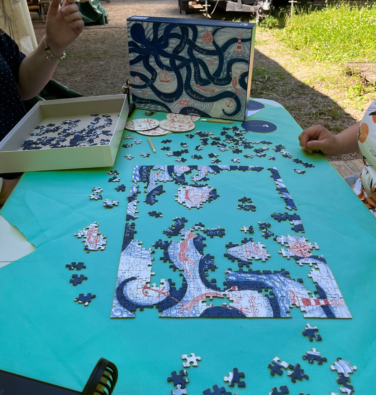 Unvollständiges Puzzle mit Oktopus-Motiv auf grünem Tuch, zwei Personen sitzen am Tisch, Puzzlebox mit Oktopusbild steht auf dem Tisch.