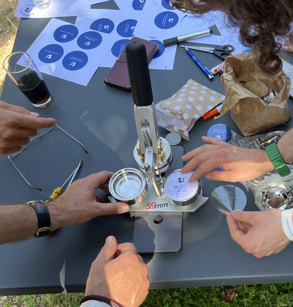 Mehrere Hände bedienen eine Button-Maschine auf einem Tisch mit runden blauen Aufklebern, Stiften und Getränken.