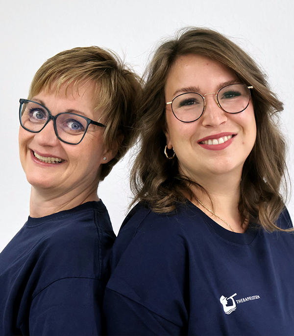 Zwei lächelnde Frauen mit Brillen stehen Rücken an Rücken, beide tragen dunkelblaue Shirts mit weißem Logo.